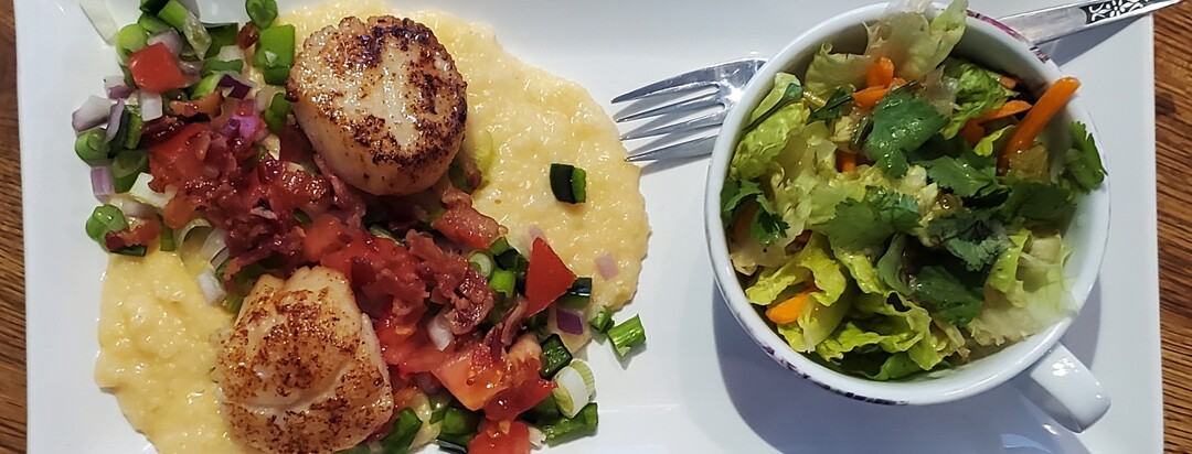 Grits garnished with bacon and scallops next to a side salad.